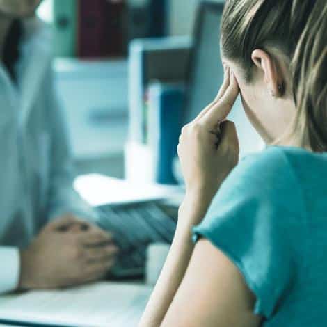 Woman suffering from bad headache or migraine. Appointment with doctor in office room. Sick and unwell lady with stress, trauma or burnout. Young patient in pain holding head with hands. Medical visit