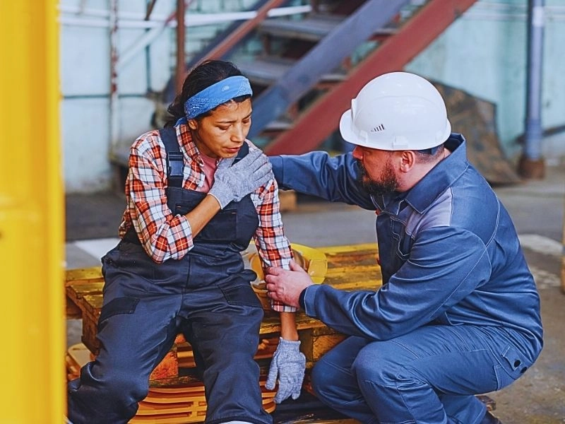 Mature foreman in workwear giving first aid to female subordinate in pain. workers' compensation shoulder injury settlements