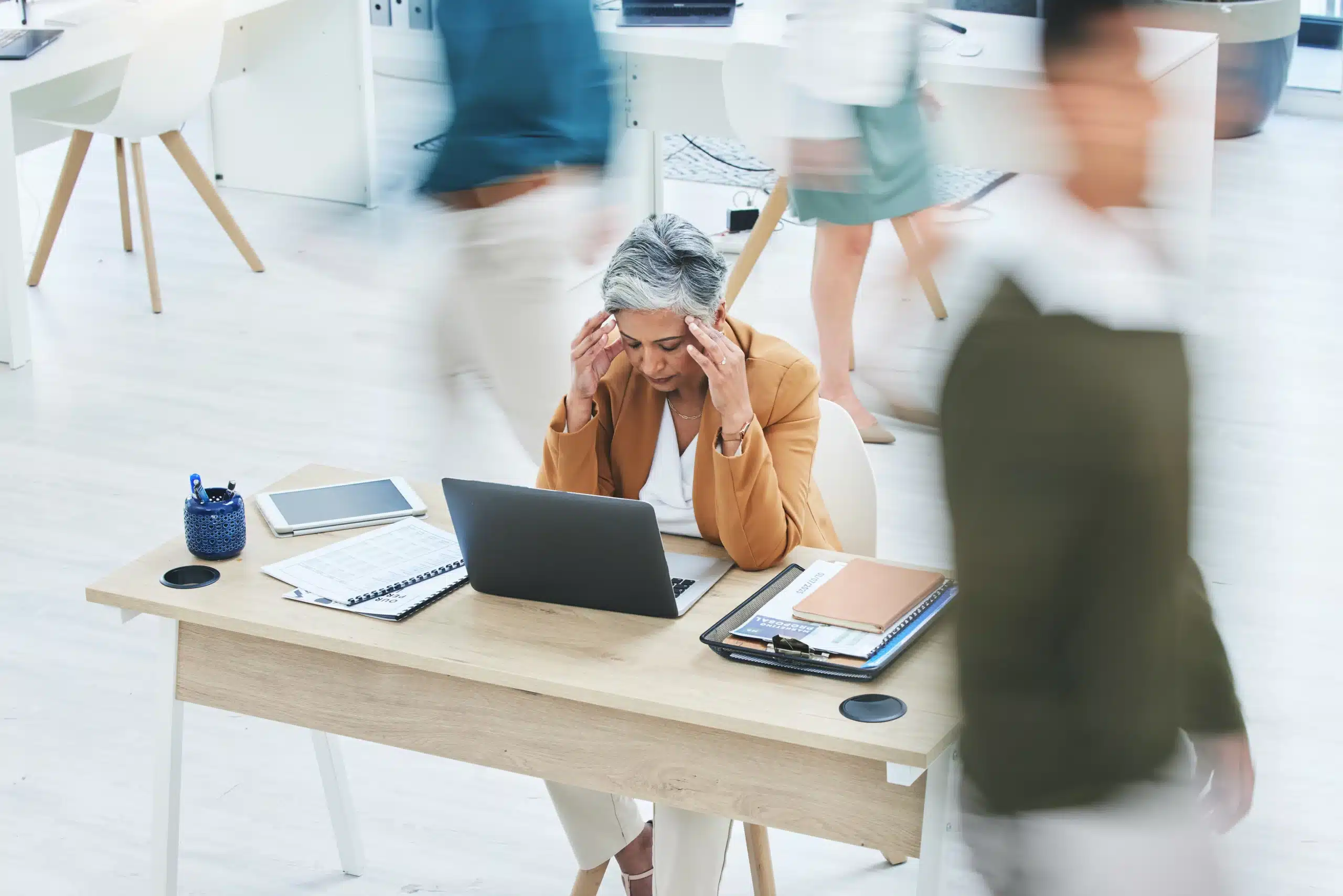 Filing a Workers’ Compensation Claim for Stress-Related Injuries in Illinois 1 A stressed woman in a brown blazer sitting at her desk with hands on her temples as blurred figures rush past in a busy office, representing workplace stress and workers' compensation claims in Illinois.