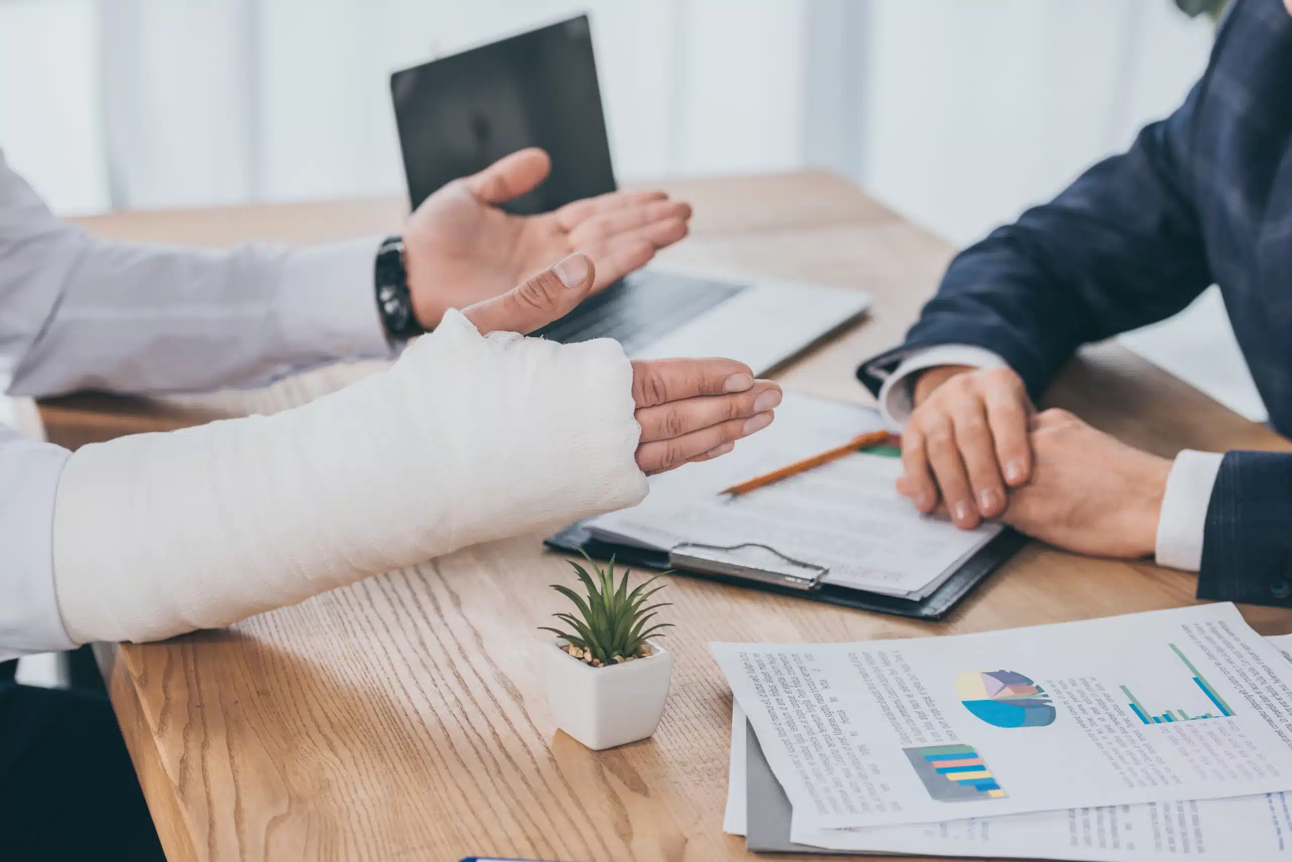 What to Expect During a Workers’ Compensation Hearing in Illinois 1 Two people sitting at a wooden desk discussing a workers' compensation case, with one person having an arm in a cast. Papers and graphs are on the table.