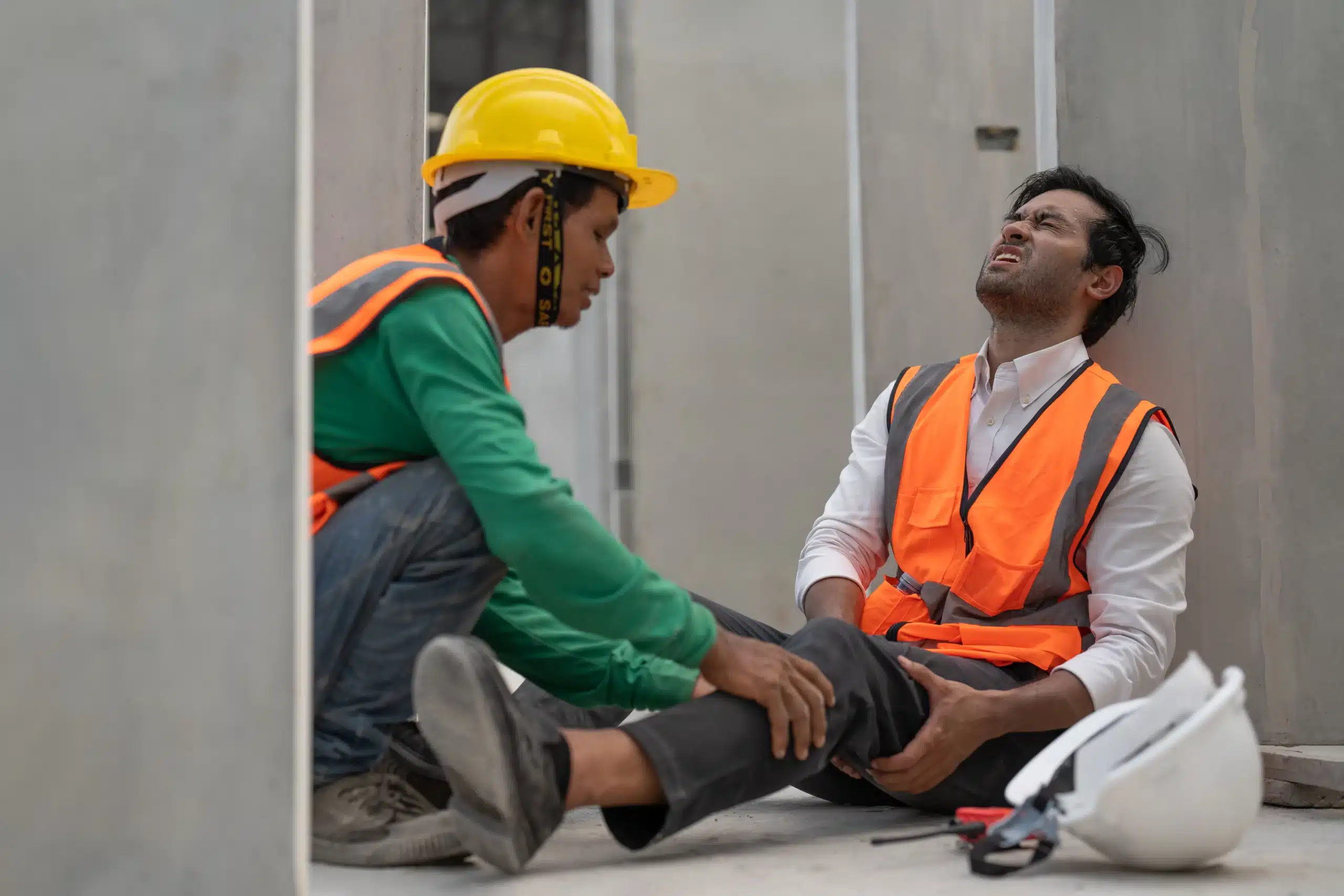 Construction worker assisting injured coworker clutching leg near job site wall