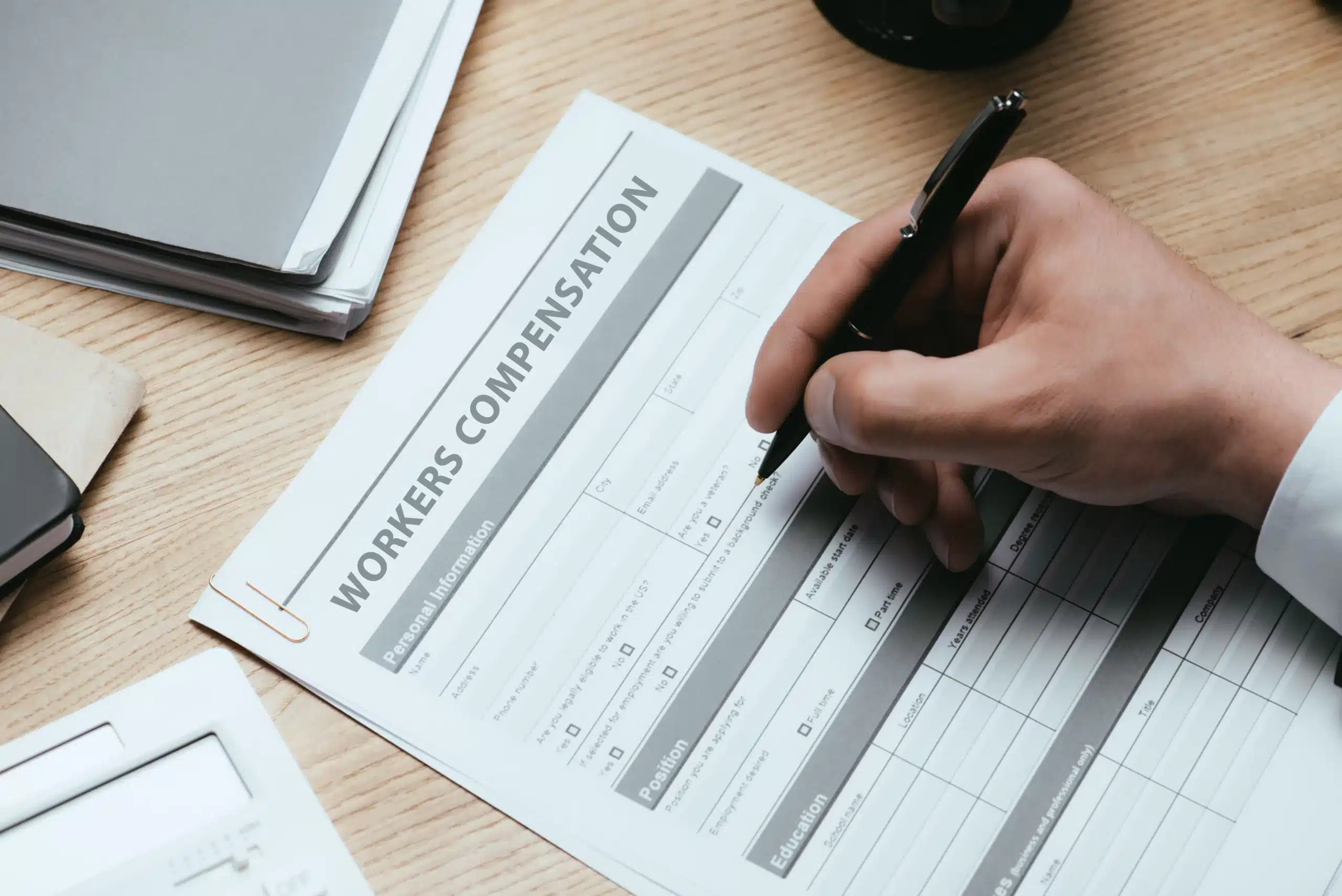 Close-up of hand filling out a workers’ compensation form with a pen on a wooden desk.