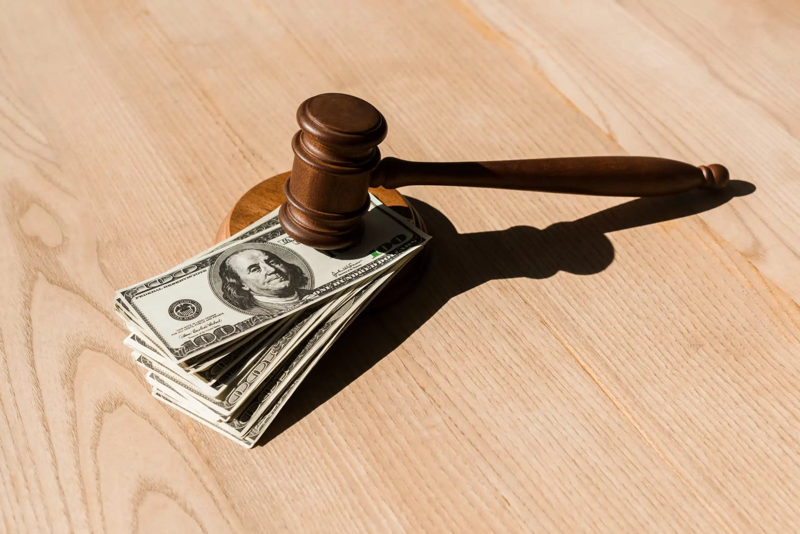 Wooden gavel on stack of hundred-dollar bills on light wood table, symbolizing legal and financial impact of medical malpractice settlements.