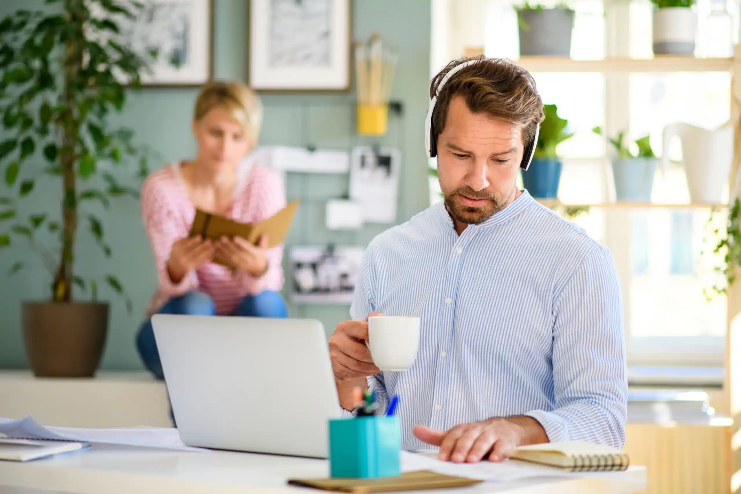 Person in headphones holding a mug and looking at a notebook on a desk with a laptop, while a woman reads in the background in a plant-filled home office, suggesting remote work and focus on workers' compensation.