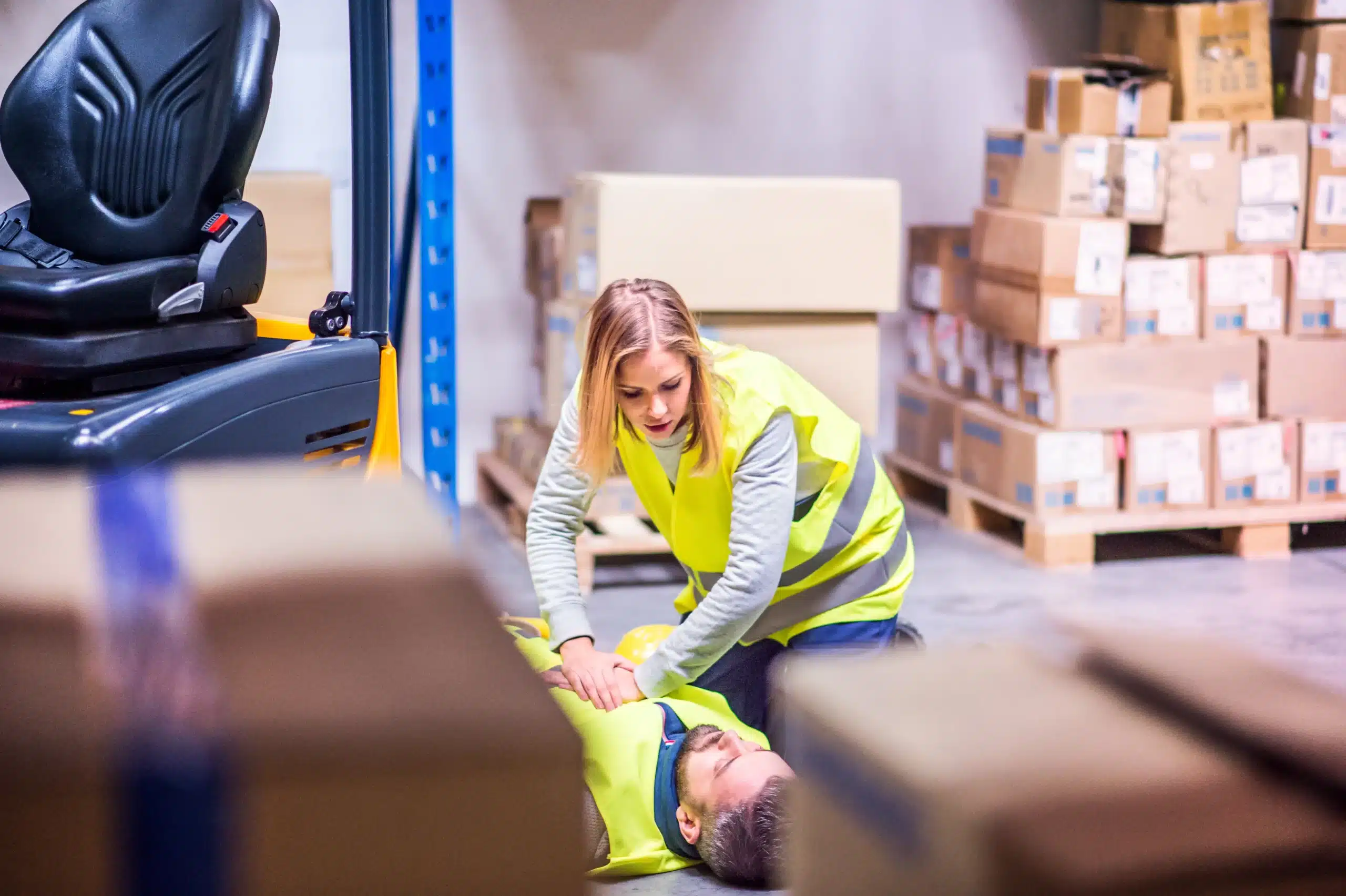 Woman in a yellow safety vest performing CPR on a man lying on a warehouse floor, surrounded by boxes and a forklift — highlighting urgency, workplace danger, and potential work‑related injury or death under workers’ compensation law.