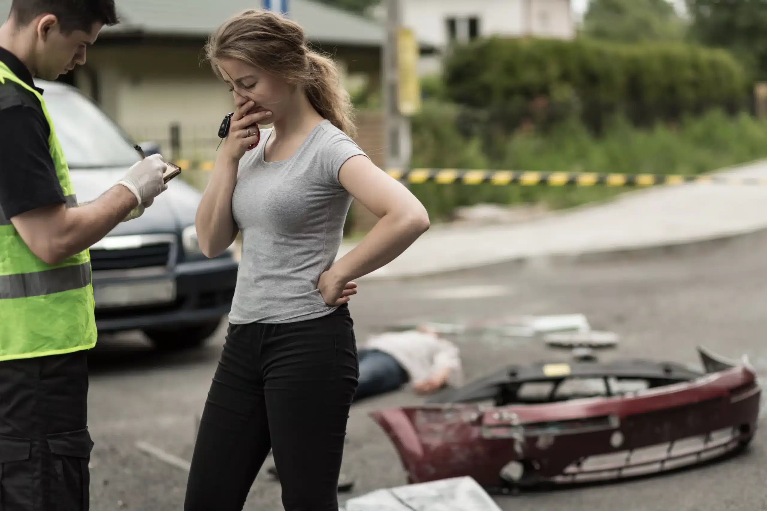 Concerned woman standing next to a police officer who is taking notes at a car accident scene, with debris and a casualty visible on the ground — illustrating serious legal consequences, car‑crash liability and comparative negligence issues.