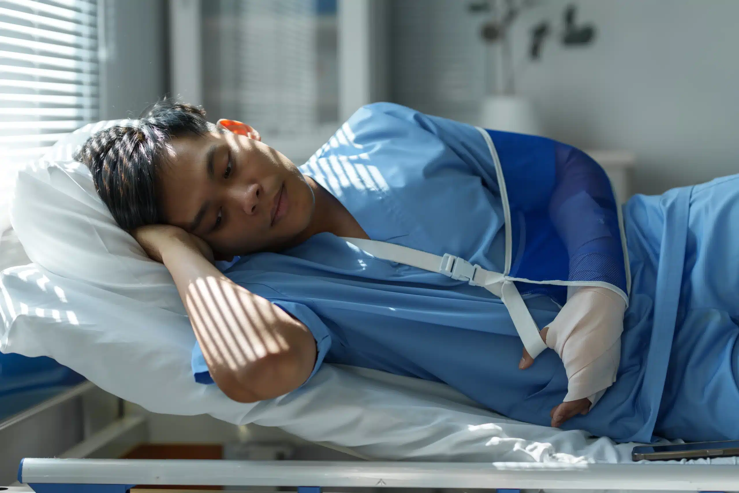 Patient in blue scrubs lying in hospital bed with arm in cast and sling, illustrating medical injury or treatment in a healthcare setting.